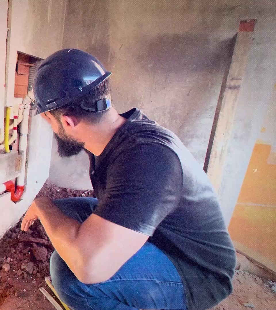 Igor Ferreto, de capacete, agachado na obra mexendo na tubulação de água embutida na parede