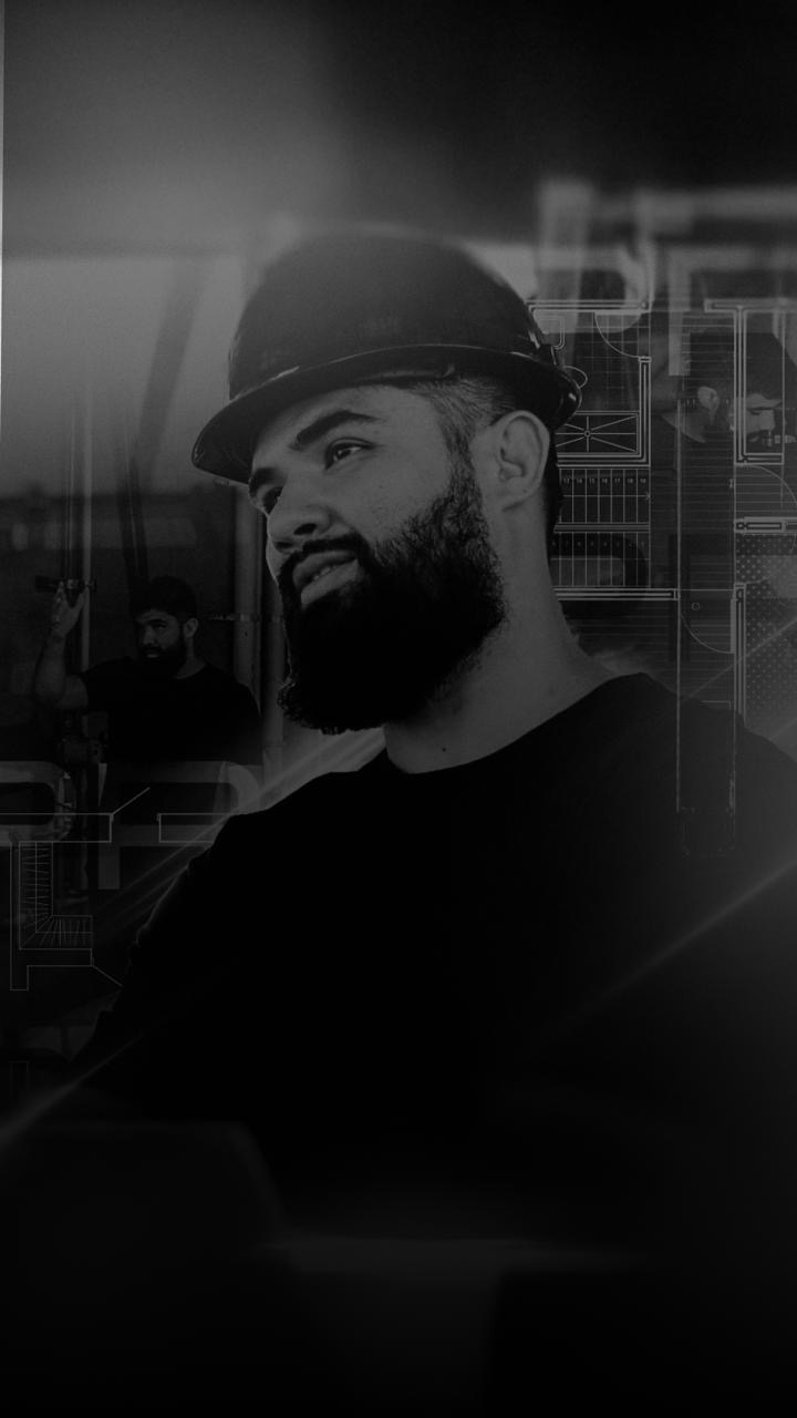 Igor Ferreto, de capacete, em retrato em preto e branco com plantas de projeto hidráulico ao fundo